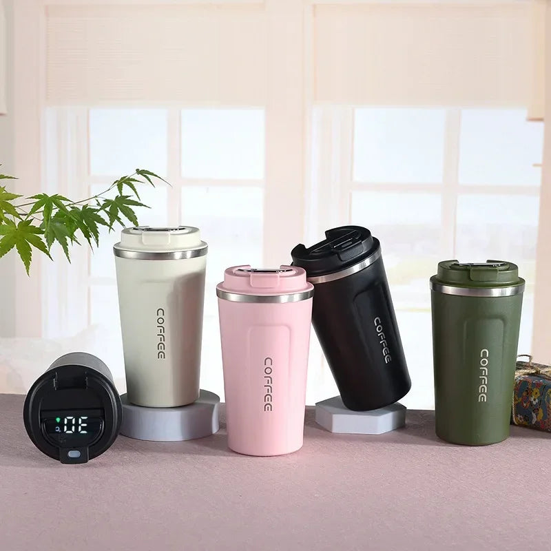 Four insulated coffee cups in different colors on a surface with a plant and digital device in the background.